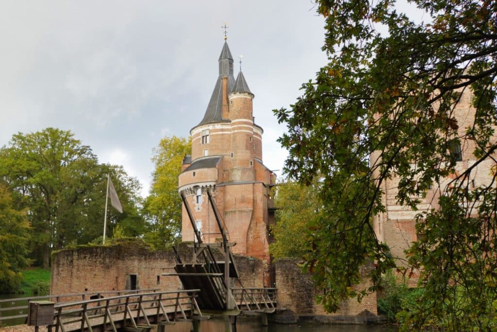Ein hoher, runder Backsteinturm mit spitzem Dach ragt hinter einer hölzernen Zugbrücke und den Ruinenmauern der Burg Wijk bij Duurstede empor, umgeben von herbstlichen Bäumen.
