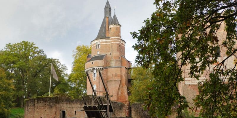 Ein hoher, runder Backsteinturm mit spitzem Dach ragt hinter einer hölzernen Zugbrücke und den Ruinenmauern der Burg Wijk bij Duurstede empor, umgeben von herbstlichen Bäumen.