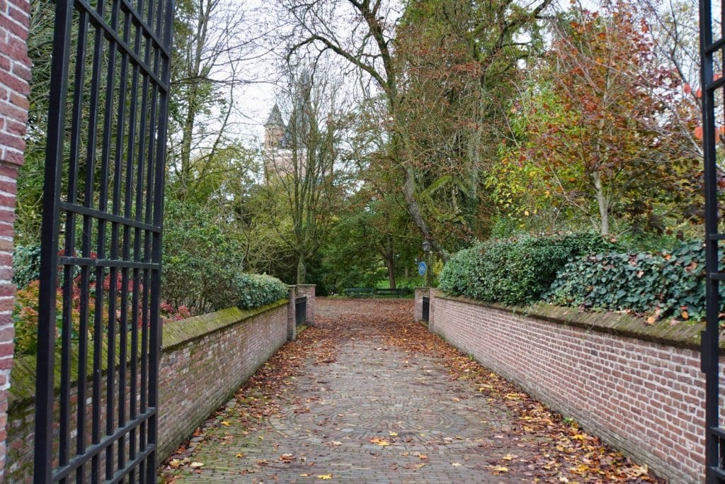 Ein gepflasterter, mit Herbstlaub bedeckter Weg führt durch ein geöffnetes schmiedeeisernes Tor in den Park von Wijk bij Duurstede, flankiert von niedrigen Backsteinmauern und dichten Bäumen.