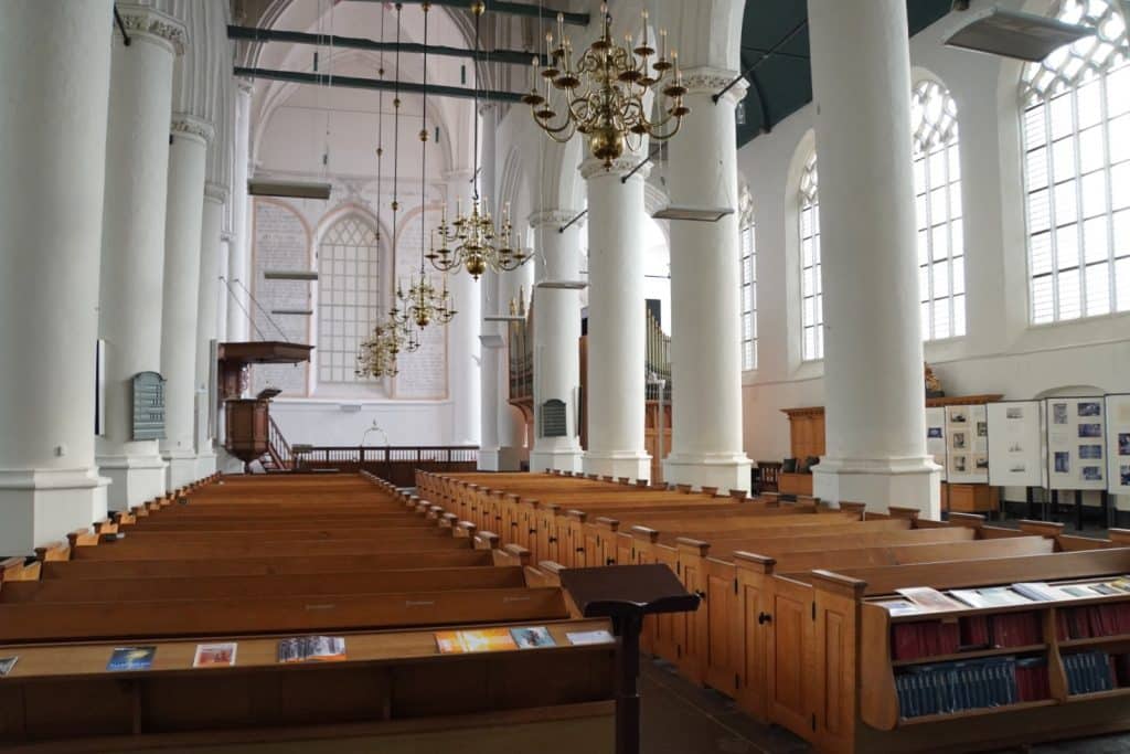 Das helle Innere der Grote Kerk in Wijk bij Duurstede zeigt lange Reihen hölzerner Kirchenbänke unter hohen weißen Säulen und prachtvollen Kronleuchtern. Im Hintergrund sind eine erhöhte Kanzel sowie große gotische Fenster zu sehen, die den weiten Raum mit natürlichem Licht fluten.