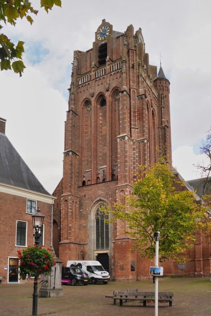 Der massive, spätgotische Backsteinturm der Grote Kerk in Wijk bij Duurstede ragt hinter einem herbstlichen Baum und einem gepflasterten Platz mit parkenden Transportern empor.links daneben steht ein historisches Backsteingebäude mit einer gusseisernen Straßenlaterne und einem Blumenkorb im Vordergrund.