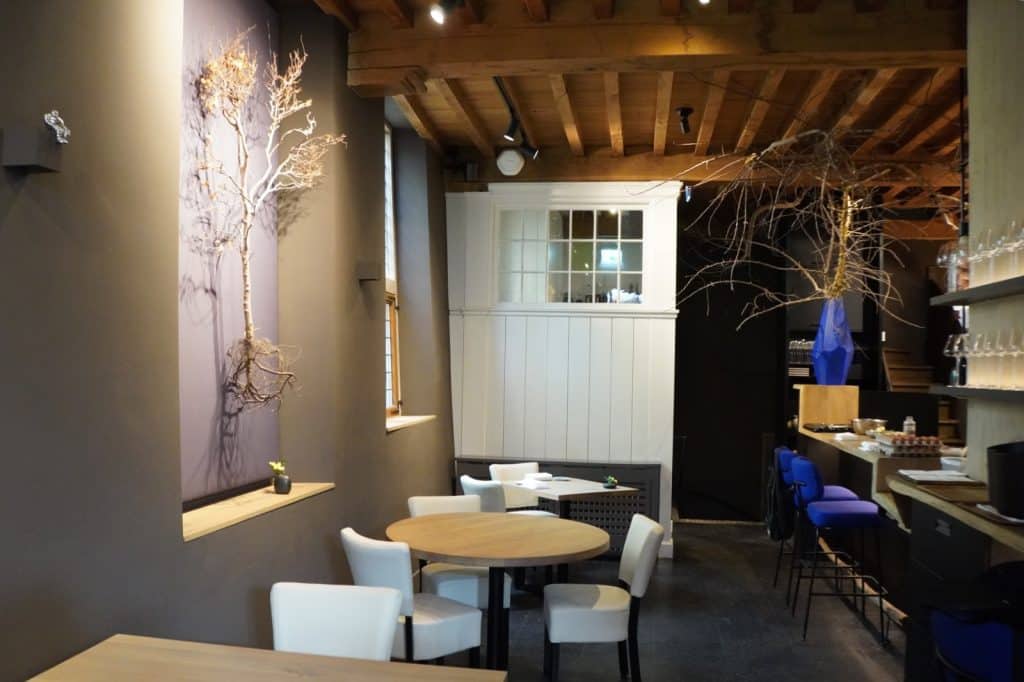 Das Restaurant Ludum in Wijk bij Duurstede zeigt einen modern gestalteten Innenraum mit Holzbalkendecke, runden Tischen mit weißen Stühlen und einer kunstvoll an der Wand befestigten, getrockneten Pflanze. Im Hintergrund ist ein Barbereich mit blauen Hockern und einer weiteren hängenden Pflanzendekoration zu sehen.