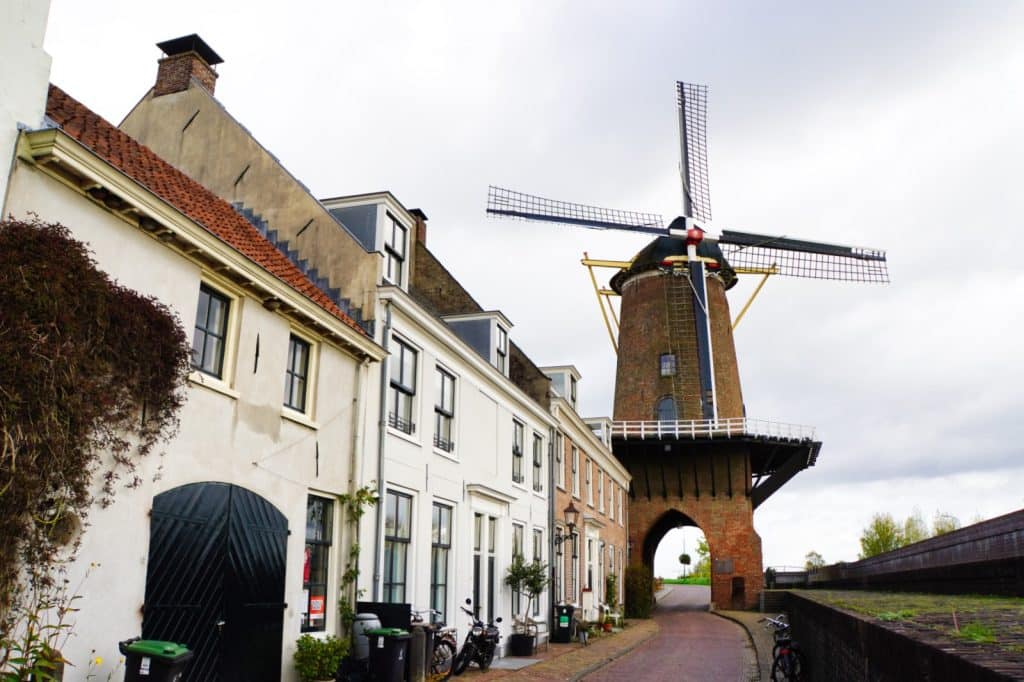 Eine schmale Straße führt unter dem Torbogen der historischen Windmühle Rijn en Lek hindurch, flankiert von einer Reihe weißer und backsteinerner Wohnhäuser.