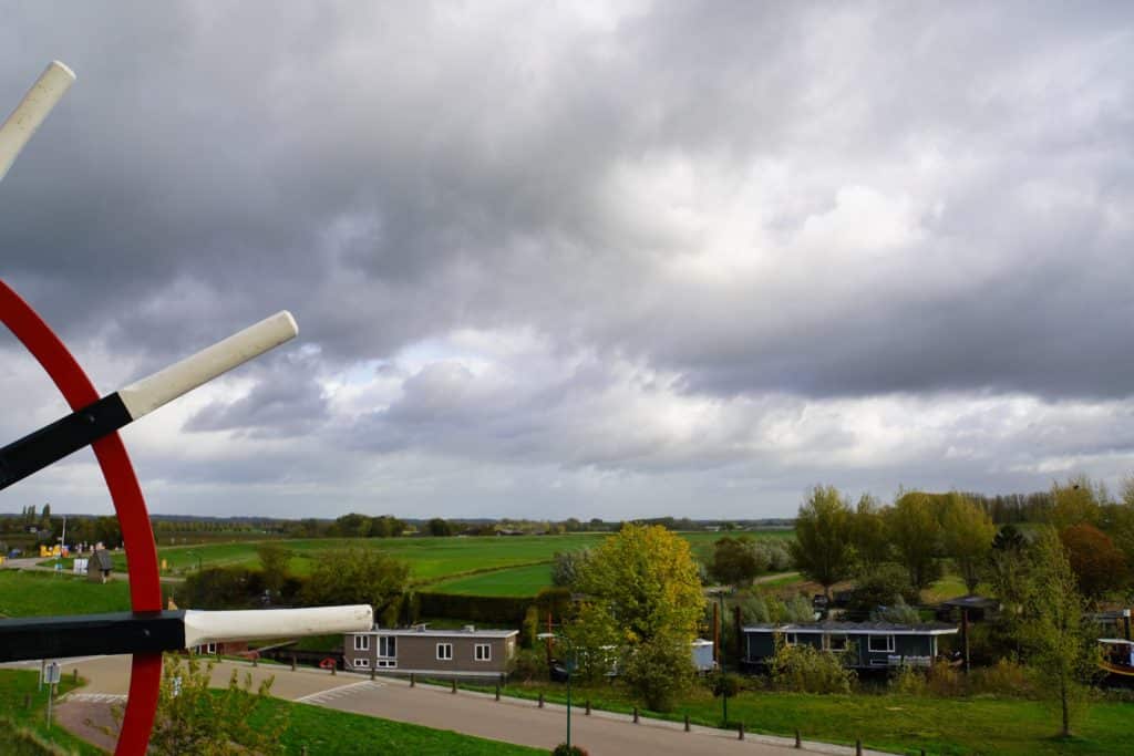 Blick von einer Mühle in Wijk bij Duurstede über eine grüne Landschaft mit Hausbooten am Ufer unter einem bewölkten Himmel, wobei im Vordergrund Teile des hölzernen Mühlenrads zu sehen sind.