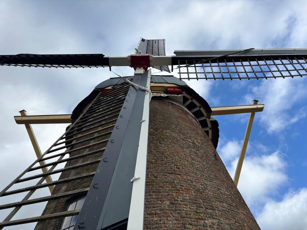 Nahaufnahme aus der Untersicht auf die historische Windmühle in Wijk bij Duurstede mit ihrem gemauerten Backsteinturm und den markanten Flügeln vor einem bewölkten Himmel.
