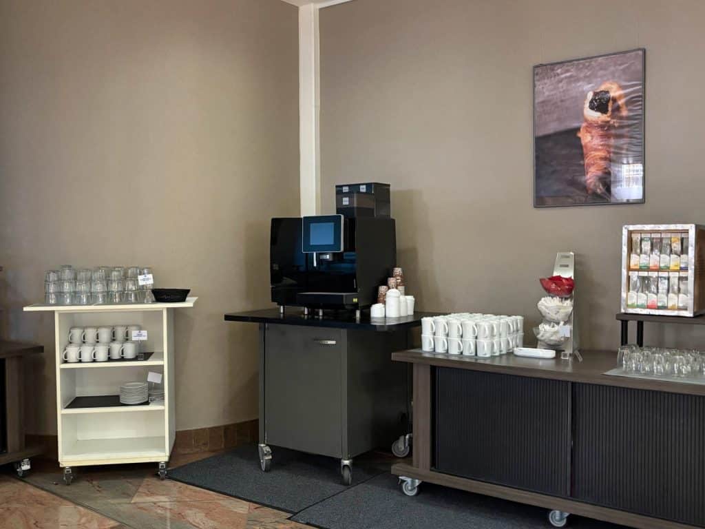 Eine Selbstbedienungs-Frühstücksstation im Maritim Hotel Hannover mit einer Kaffeemaschine auf einem Rollwagen, flankiert von Regalen mit Tassen, Gläsern und einer Auswahl an Teebeuteln.