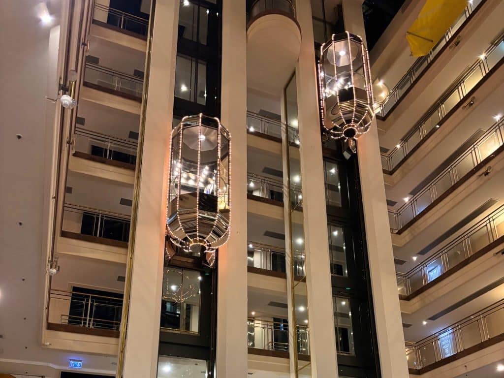 Blick in das Atrium des Maritim Hotels Hannover mit zwei beleuchteten, gläsernen Panoramaaufzügen, die vor den umlaufenden Balkonbrüstungen der Etagen emporfahren.
