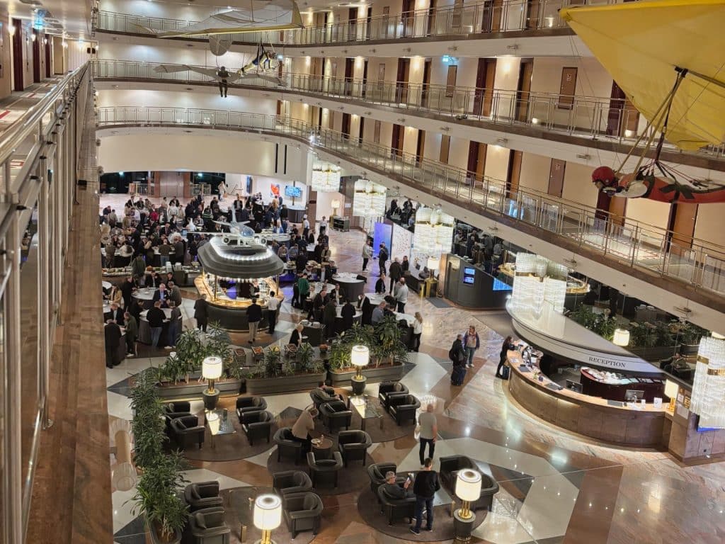 Blick von oben in die weitläufige, mehrstöckige Lobby des Maritim Airport Hotels Hannover mit zahlreichen Gästen, Sitzgruppen, einer Rezeption und dekorativen Fluggeräten, die unter der Decke hängen.