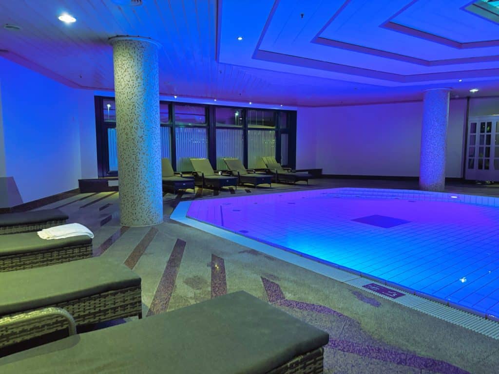 Der Innenpool des Maritim Hotels Hannover ist in stimmungsvolles blaues Licht getaucht und von bequemen Liegestühlen auf einem gemusterten Steinboden umgeben.