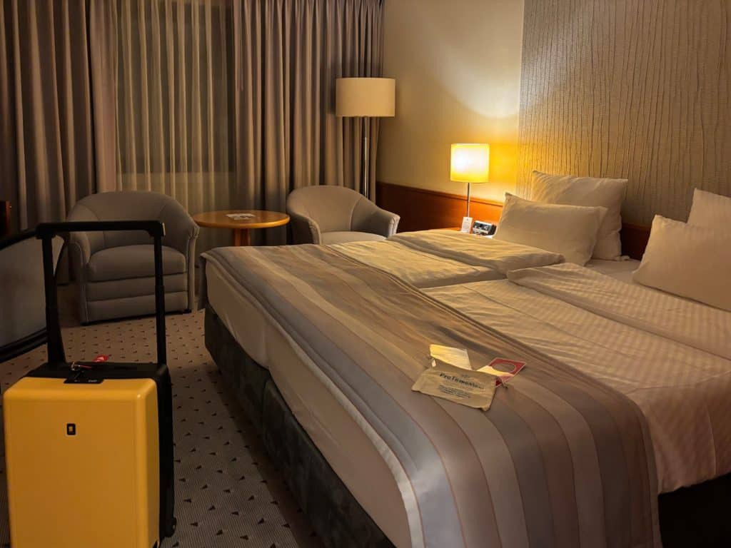 Ein gemütliches Hotelzimmer im Maritim Hotel Hannover mit einem großen Doppelbett, zwei grauen Sesseln, einem kleinen runden Tisch und einem gelben Rollkoffer im Vordergrund.