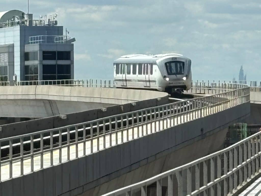 Ein weißer New York AirTrain JFK fährt auf einer erhöhten Betonkurve an einem modernen Bahnhofsgebäude vorbei, während im Hintergrund die Skyline der Stadt unter einem bewölkten Himmel zu sehen ist.