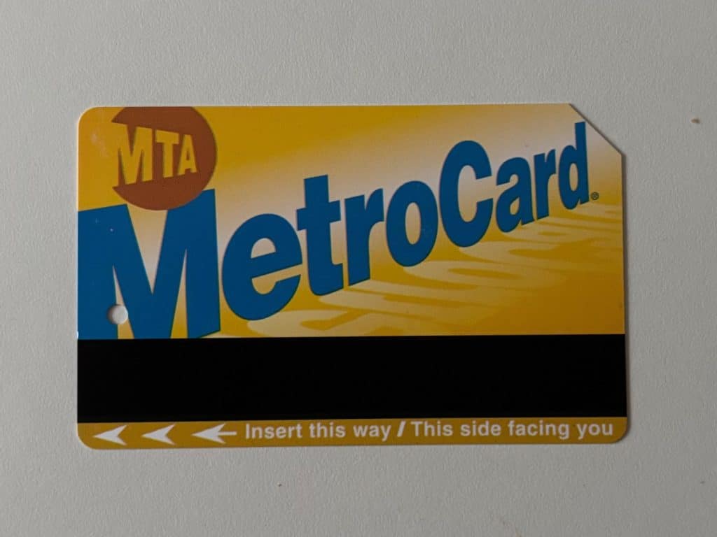 Eine gelbe New Yorker MetroCard mit blauem Schriftzug und schwarzem Magnetstreifen. Am unteren Rand befinden sich weiße Pfeile und der Text „Insert this way / This side facing you“.