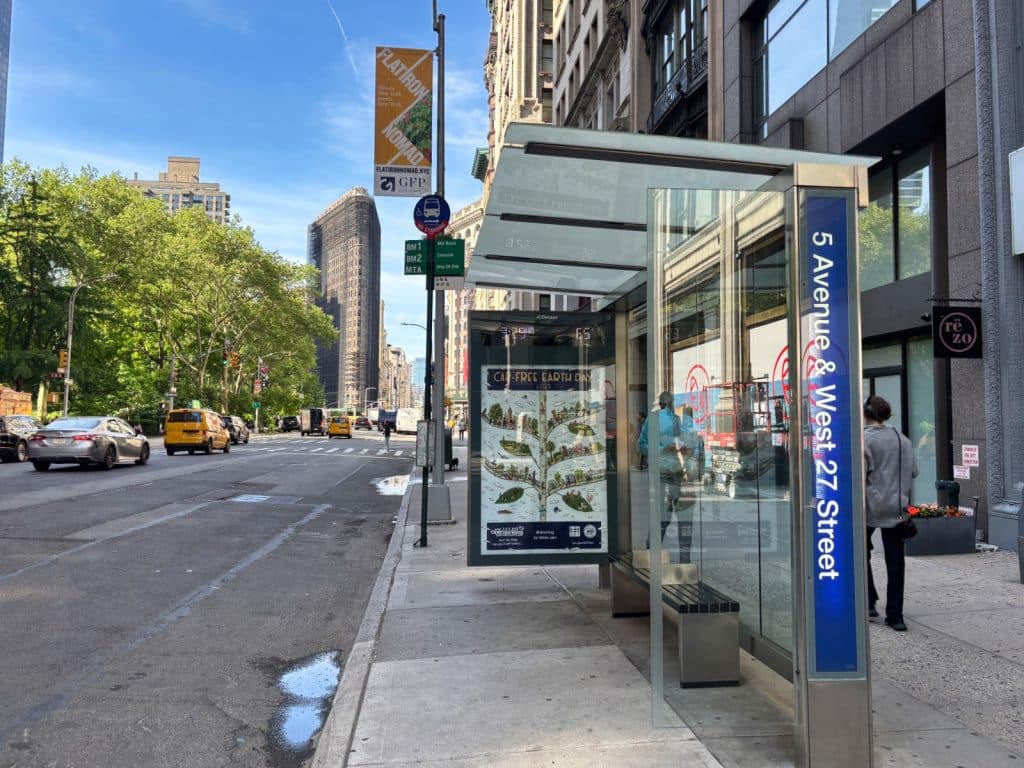 Eine moderne Bushaltestelle in New York an der Ecke 5th Avenue und West 27th Street mit einer gläsernen Wartehalle, Werbeplakaten und Blick auf das belebte Straßenbild mit dem Flatiron Building im Hintergrund.