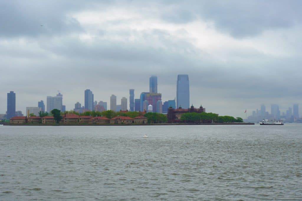 Blick über das Wasser auf Ellis Island in New York mit seinen historischen Backsteingebäuden vor der nebligen Skyline von Jersey City unter einem wolkenverhangenen Himmel.