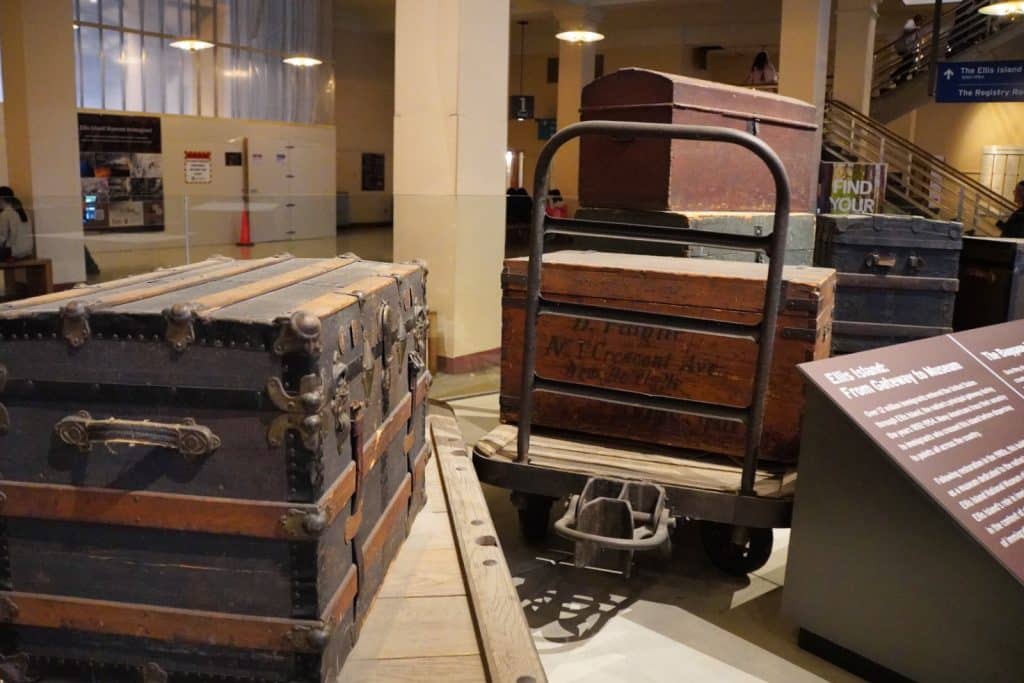 Historische Reisekoffer und Truhen aus Holz und Metall sind im Gepäckraum des Ellis Island National Museum of Immigration in New York auf einem hölzernen Gepäckwagen und dem Boden ausgestellt.