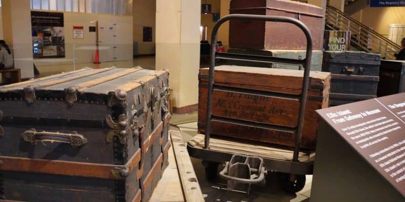 Historische Reisekoffer und Truhen aus Holz und Metall sind im Gepäckraum des Ellis Island National Museum of Immigration in New York auf einem hölzernen Gepäckwagen und dem Boden ausgestellt.
