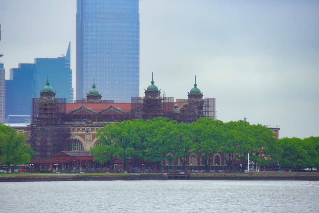 Das Hauptgebäude von Ellis Island in New York mit seinen markanten vier Türmen und grünen Kuppeln steht hinter einer Baumreihe am Wasser, während im Hintergrund moderne Wolkenkratzer aufragen.