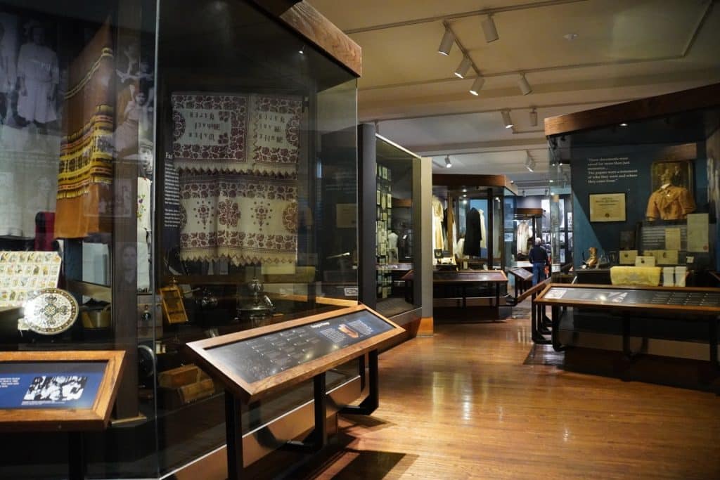Eine Innenansicht des Ellis Island Museums in New York zeigt verschiedene beleuchtete Glasvitrinen mit historischen Exponaten wie Textilien, Kleidung und Dokumenten auf einem glänzenden Holzboden. Informationstafeln auf schrägen Pulten stehen vor den Ausstellungen in dem weitläufigen, musealen Raum.