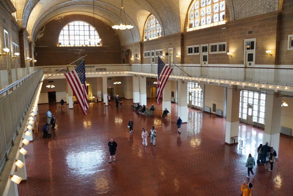 Blick von oben in die weitläufige Registrierungshalle von Ellis Island in New York, die mit einem glänzenden roten Fliesenboden, hohen Rundbogenfenstern und zwei großen, von der Empore hängenden US-Flaggen ausgestattet ist.