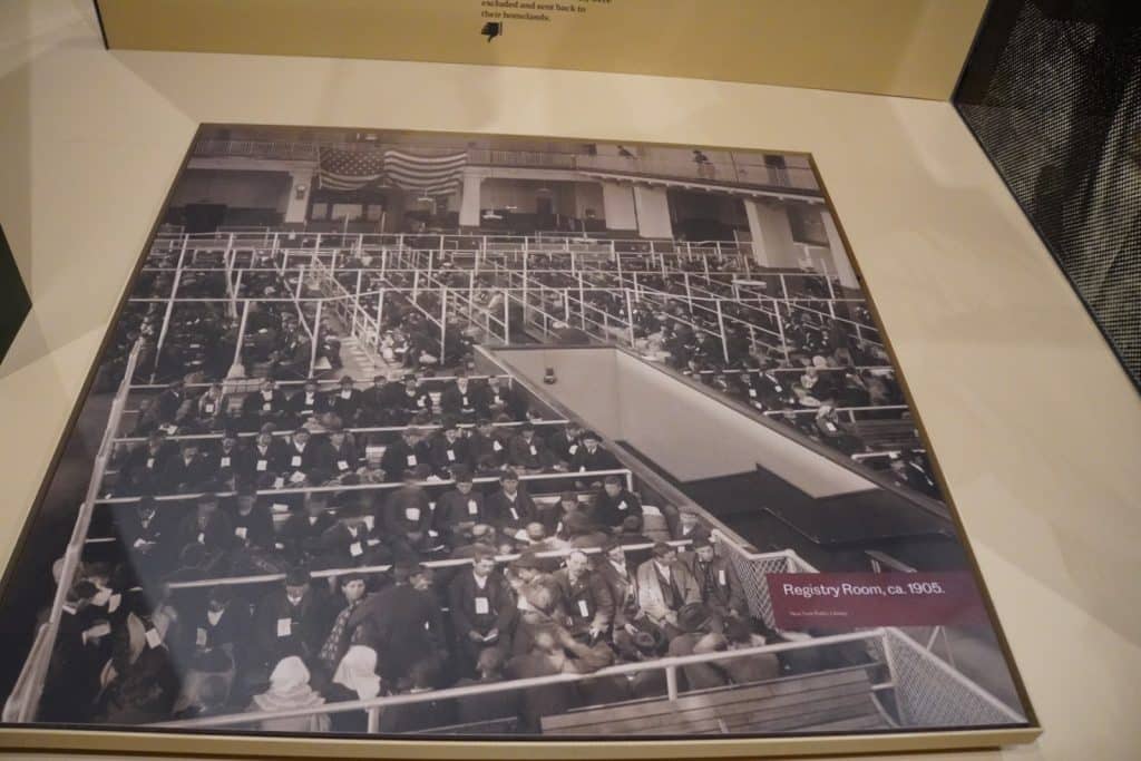Eine historische Schwarz-Weiß-Fotografie der Registrierungshalle auf Ellis Island in New York um 1905 zeigt zahlreiche Einwanderer, die in langen, durch Metallgitter abgetrennten Sitzreihen auf ihre Bearbeitung warten. Im Hintergrund hängen amerikanische Flaggen von der Empore des großen, hellen Saals herab.