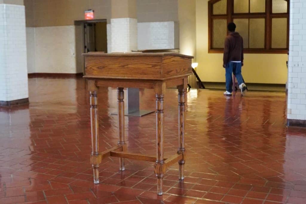 Ein hölzernes Registrierungspult steht im Vordergrund auf dem glänzenden Fliesenboden eines Gebäudes auf Ellis Island in New York, während im Hintergrund eine Person an einem Fenster vorbeigeht.