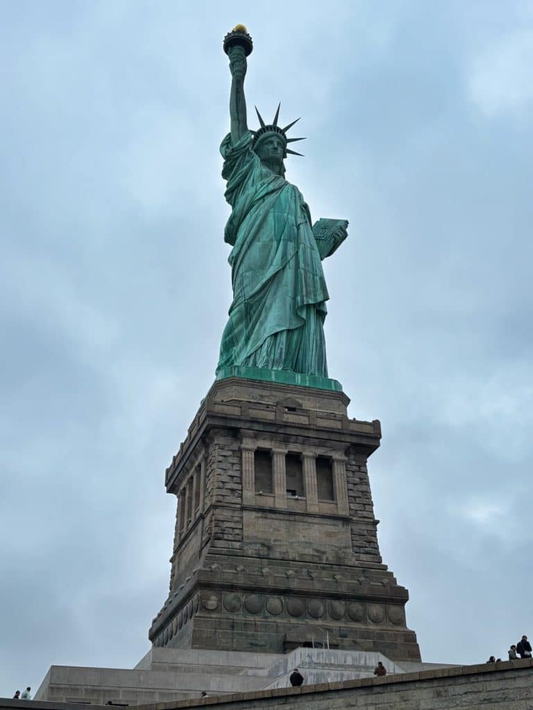 Die Freiheitsstatue in New York ragt auf ihrem massiven Steinsockel vor einem bewölkten, grauen Himmel empor, während sie die Fackel hochhält und eine Gesetzestafel im Arm trägt.
