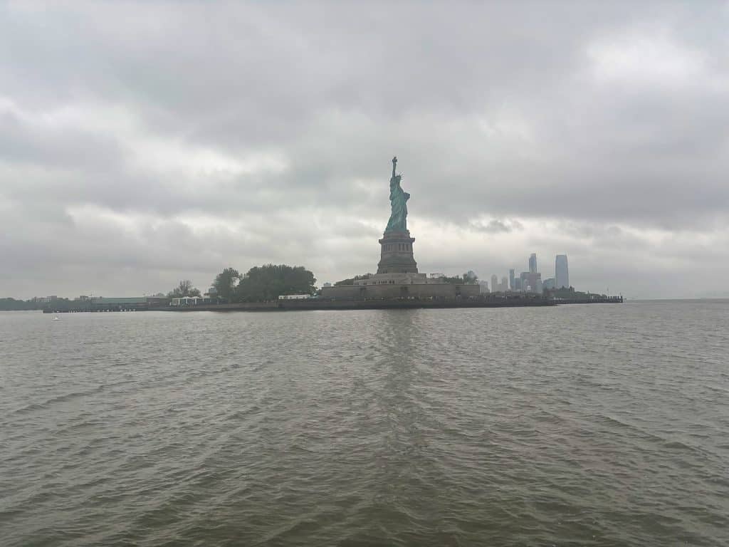 Die Freiheitsstatue in New York steht auf Liberty Island unter einem wolkenverhangenen, grauen Himmel, während das Wasser im Vordergrund leichte Wellen schlägt. Im Hintergrund sind schemenhaft die Hochhäuser der Skyline bei regnerischem Wetter zu erkennen.