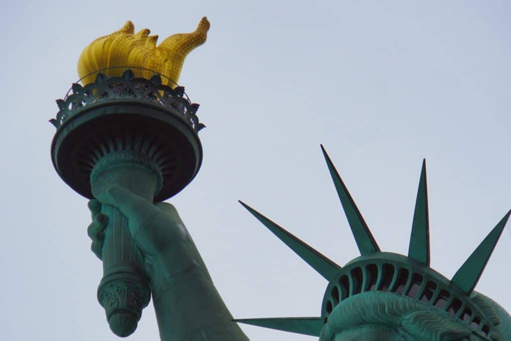 Nahaufnahme der Freiheitsstatue in New York, die den erhobenen Arm mit der goldfarbenen Fackel und einen Teil des Kopfes mit der gezackten Krone vor einem hellen Himmel zeigt.