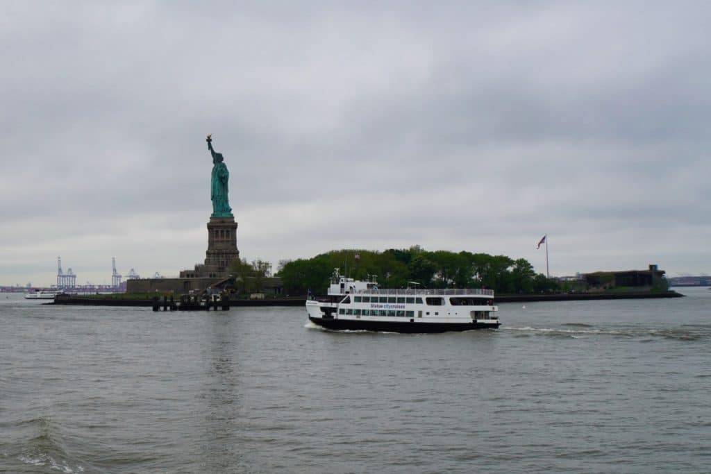Eine weiße Fähre fährt auf dem Wasser vor der Freiheitsstatue in New York unter einem bewölkten Himmel. Im Hintergrund ist die bewaldete Insel mit dem Denkmal und Hafenkränen am Horizont zu sehen.
