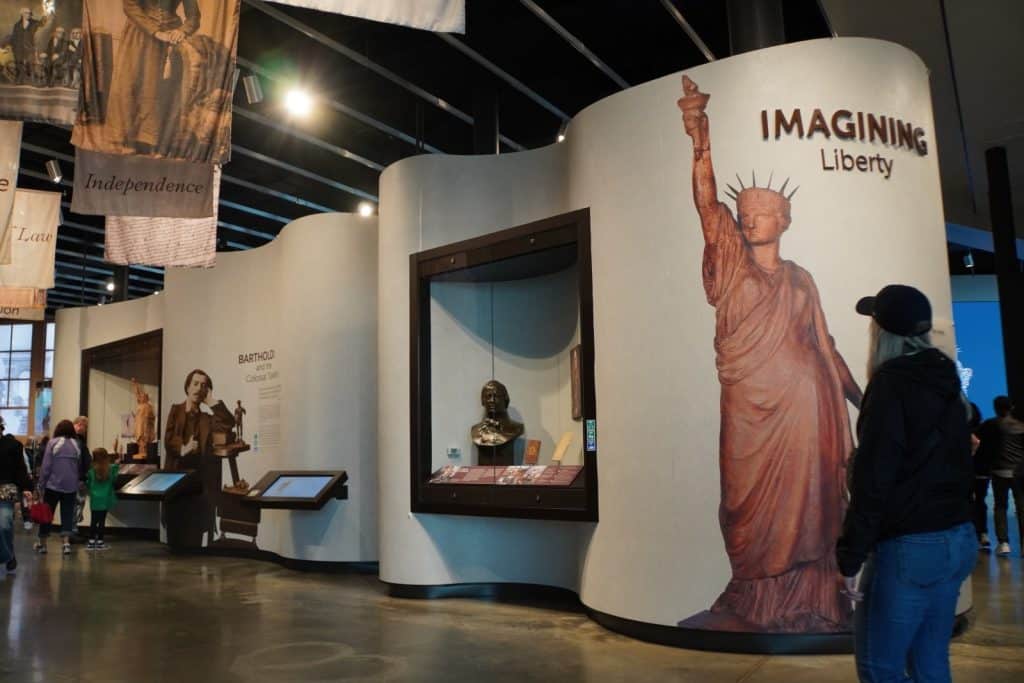 Besucher betrachten im Museum der Freiheitsstatue in New York eine Ausstellung mit dem Titel „Imagining Liberty“, die großformatige Grafiken der Statue sowie historische Büsten und Informationstafeln in geschwungenen Ausstellungswänden zeigt.