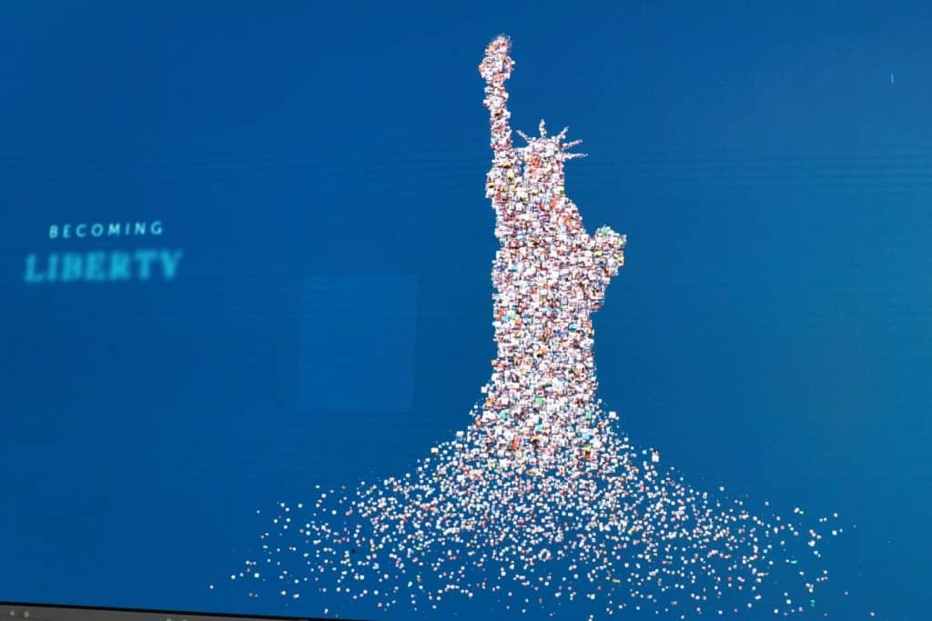 Eine digitale Fotocollage im Statue of Liberty Museum zeigt die Freiheitsstatue von New York, die aus unzähligen kleinen Einzelporträts zusammengesetzt ist, vor einem blauen Hintergrund mit dem Schriftzug „BECOMING LIBERTY“.