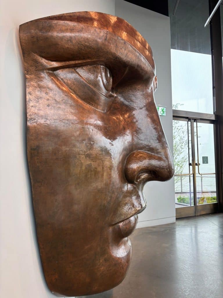 Eine Nahaufnahme im Profil zeigt eine kupferfarbene Nachbildung des Gesichts der Freiheitsstatue, die an einer weißen Wand im Statue of Liberty Museum in New York montiert ist.