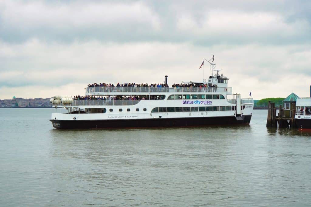 Ein großes, weißes Ausflugsschiff von Statue City Cruises, voll besetzt mit Passagieren, fährt auf dem Wasser in Richtung der New Yorker Freiheitsstatue unter einem bewölkten Himmel.
