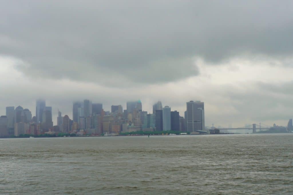Eine Weitwinkelaufnahme der Skyline von New York City unter einem wolkenverhangenen, grauen Himmel, gesehen von der Wasserseite aus. Die Wolken hängen tief über den Wolkenkratzern von Lower Manhattan, während im Hintergrund rechts die Brooklyn Bridge im Dunst erkennbar ist.