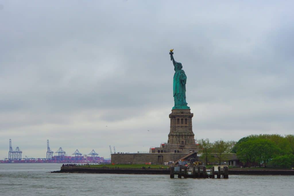 Die Freiheitsstatue in New York steht auf ihrem steinernen Sockel auf Liberty Island unter einem bewölkten Himmel, während im Hintergrund Hafenkräne am Ufer zu sehen sind.