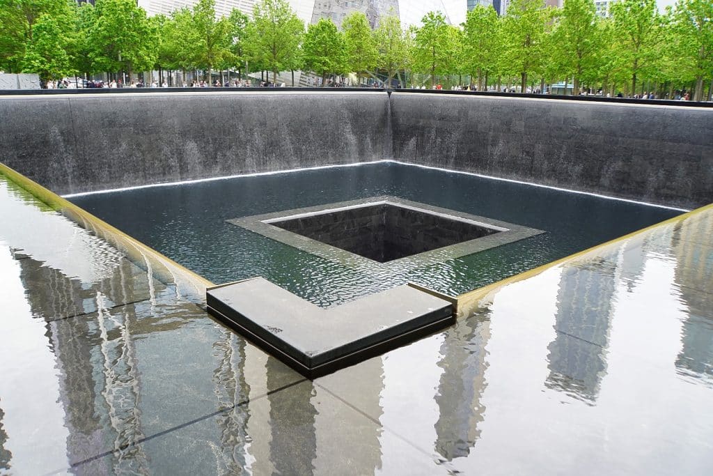 Ein quadratisches Wasserbecken am Ground Zero in New York, bei dem Wasser an den dunklen Steinwänden in die Tiefe stürzt und in einer zentralen, tiefer gelegenen Öffnung verschwindet, umgeben von grünen Bäumen.