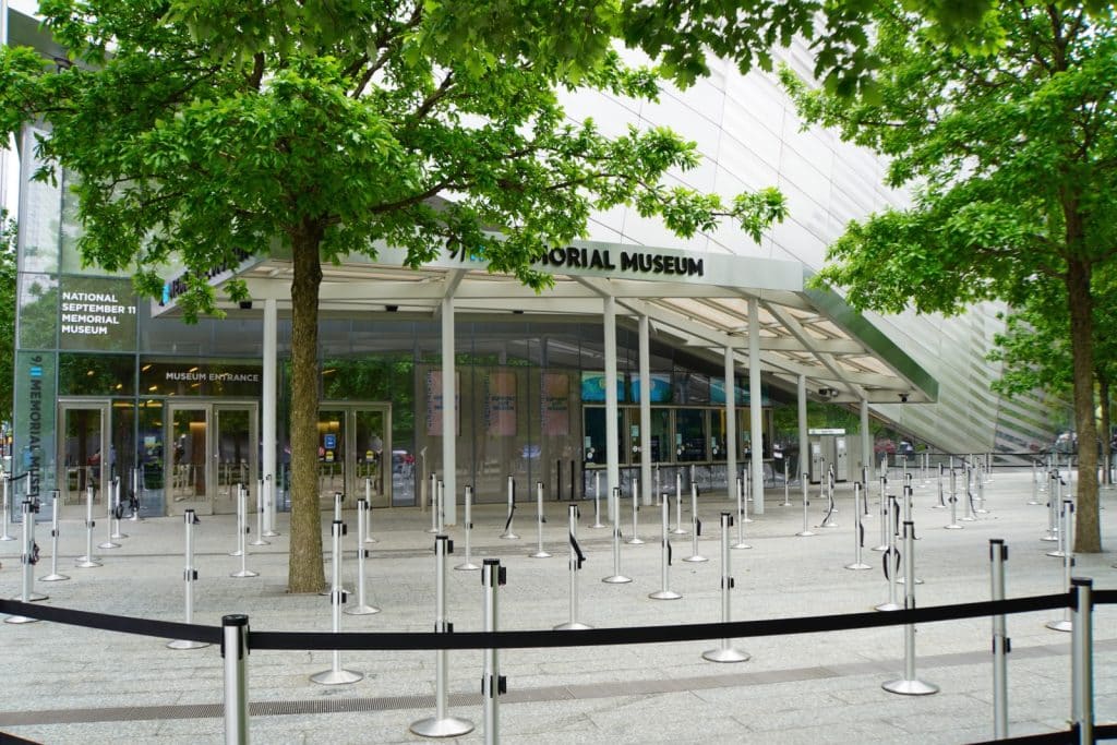 Der Eingangsbereich des National September 11 Memorial Museum am Ground Zero in New York mit einem modernen Glasgebäude, weißen Stützsäulen und zahlreichen Absperrpfosten für Warteschlangen unter grünen Bäumen.