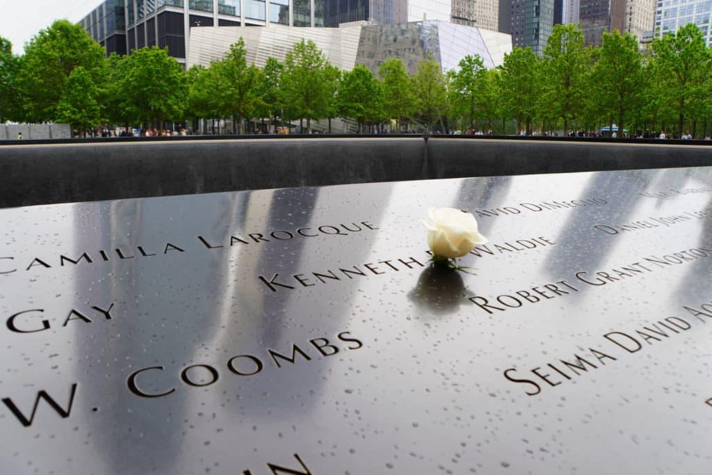 Nahaufnahme einer Gedenktafel am Ground Zero in New York mit eingravierten Namen, auf der eine einzelne weiße Rose liegt, vor dem Hintergrund von Bäumen und Gebäuden.