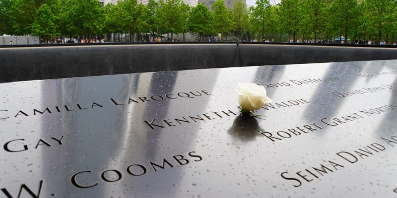 Nahaufnahme einer Gedenktafel am Ground Zero in New York mit eingravierten Namen, auf der eine einzelne weiße Rose liegt, vor dem Hintergrund von Bäumen und Gebäuden.
