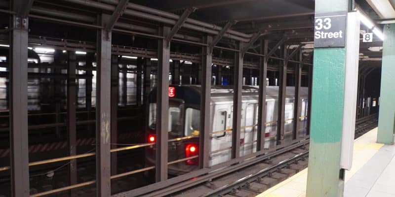 Ein New Yorker U-Bahn-Zug der Linie 4 fährt durch die Station 33rd Street, gesehen von einem Bahnsteig aus hinter stählernen Stützpfeilern.