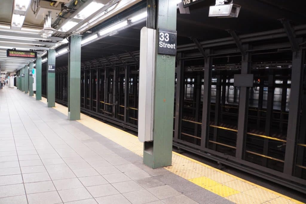 Ein leerer Bahnsteig der New Yorker Metro-Station 33rd Street mit grünen Säulen, hellen Fliesen und Blick auf die dunklen Gleise hinter einem Metallgitter.