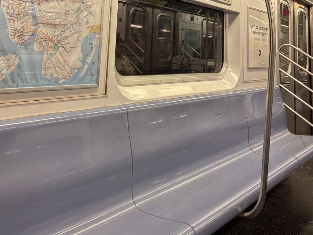 Innenansicht eines New Yorker U-Bahn-Wagens mit hellblauen Kunststoffsitzbänken, einem Streckennetzplan an der Wand und einem Fenster, in dem sich das gegenüberliegende Abteil spiegelt.