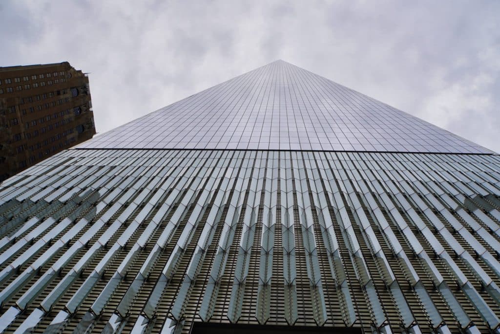 Das Foto zeigt eine Froschperspektive auf das One World Trade Center in New York. Der untere Teil des Gebäudes ist mit vertikalen, silbernen Elementen verziert, während der obere Teil eine glatte, pyramidenförmige Glasfassade aufweist, die sich gegen einen bewölkten Himmel abhebt. Links ist ein weiteres Gebäude mit einer Fassade aus braunen Ziegeln zu sehen.