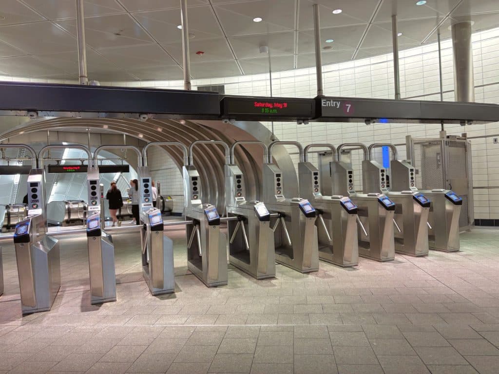 Eine Reihe moderner Edelstahl-Drehkreuze am Eingang einer New Yorker Metrostation unter einer digitalen Anzeige, die das Datum und die Uhrzeit anzeigt. Im Hintergrund sind Passanten und die gewölbte Architektur des U-Bahnhofs zu sehen.