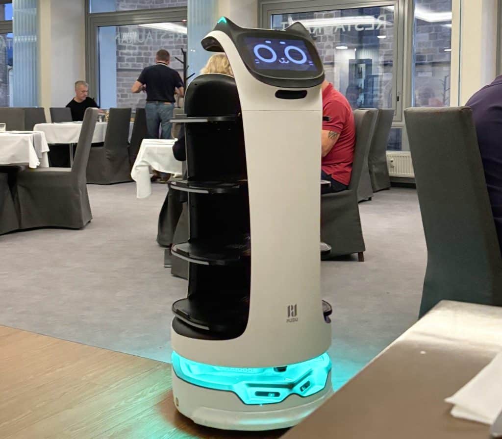 Ein weißer Servierroboter namens Robbi mit Katzenohren, einem digitalen Katzengesicht und leuchtend blauer Basis steht im Restaurant des Hotel Central in Pilsen. Er besitzt mehrere offene Ablagefächer für Speisen und Getränke und befindet sich inmitten eines modern eingerichteten Gastraums mit Gästen.