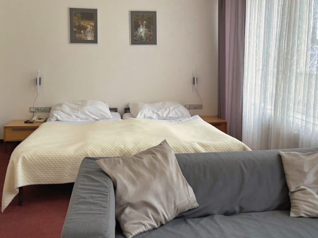 Ein helles Hotelzimmer im Hotel Central in Pilsen mit einem Doppelbett, das mit einer cremefarbenen Tagesdecke bezogen ist, und einem grauen Sofa mit Kissen im Vordergrund. Über dem Bett hängen zwei gerahmte Bilder an der weißen Wand neben einem Fenster mit hellen, transparenten Vorhängen.