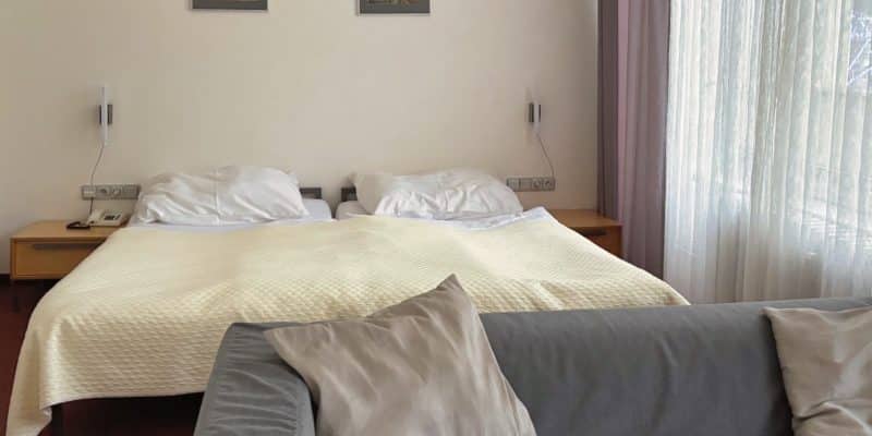 Ein helles Hotelzimmer im Hotel Central in Pilsen mit einem Doppelbett, das mit einer cremefarbenen Tagesdecke bezogen ist, und einem grauen Sofa mit Kissen im Vordergrund. Über dem Bett hängen zwei gerahmte Bilder an der weißen Wand neben einem Fenster mit hellen, transparenten Vorhängen.