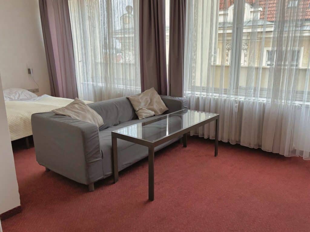 Ein Hotelzimmer im Hotel Central in Pilsen mit einem grauen Sofa, einem Glastisch und einem Bett auf rotem Teppichboden vor großen Fenstern mit hellen Vorhängen.