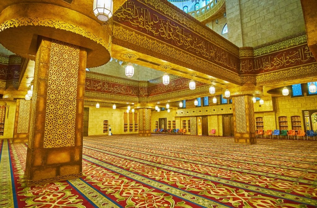 Das prachtvolle Innere der Al-Sahaba-Moschee in Sharm El Sheikh zeigt goldene Säulen mit geometrischen Mustern, kunstvolle arabische Inschriften an den Deckenbalken und einen weitläufigen, rot gemusterten Gebetsteppich. Zahlreiche hängende Laternen beleuchten den sakralen Raum, während im Hintergrund farbenfrohe Stühle an den Wänden aufgereiht sind.