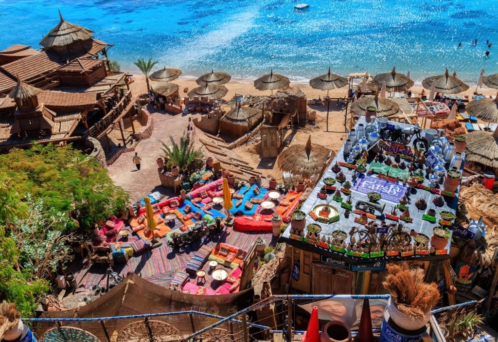 Blick von oben auf das farbenfrohe Farsha Café in Sharm El Sheikh, das mit bunten Sitzkissen, Teppichen und traditioneller Dekoration an einem Felshang direkt über dem türkisfarbenen Meer liegt.